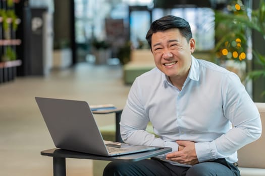 Disappointed sick Chinese businessman feeling overeating during work process on laptop computer in office lobby. Upset ill male manager guy suffering from painful stomach ache period cramps, poisoning