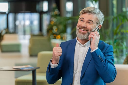 Happy mature businessman in suit answering to client on mobile phone call, using smartphone and laptop. Conversation. Caucasian bearded man guy working online job, talking in modern office lobby.