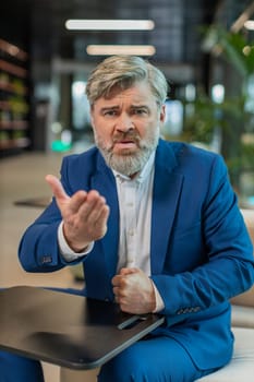Conflict Quarrel Negotiation. Mature angry boss businessman making webcam online video call, talking colleague conversation looking at camera. Caucasian guy in suit sitting in modern office lobby.