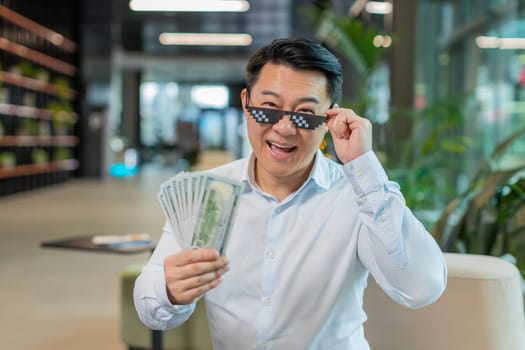 Happy rich Chinese mature businessman putting on thug life sunglasses showing money dollar cash banknotes sitting in modern office lobby. Success business career, lottery winner, big income, wealth.