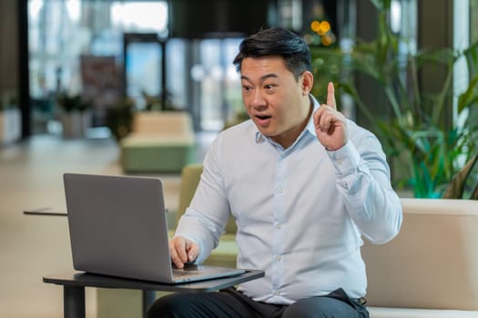 Eureka. Thoughtful clever inspired Chinese businessman using laptop makes gesture raises finger came up with creative plan feels excited with good idea inspiration motivation question in hotel lobby.