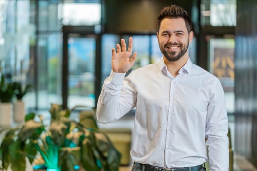 Hello. Happy young Caucasian businessman employee waves hand hi gesture greeting welcomes someone, online webinar, remote distance conversation. Entrepreneur man in shirt standing in office lobby.