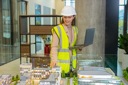 Confident young female architect in hardhat and reflective jacket using laptop and analyzing business office complex architectural model. Woman girl smart designer of buildings working in office.