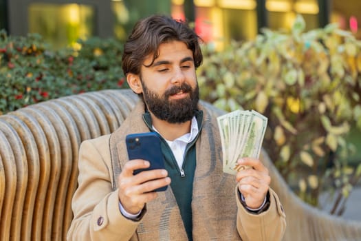Happy rich Indian businessman tourist using smartphone and counting money dollar cash banknotes sitting on bench on downtown city street. Success business career, lottery winner, big income, wealth