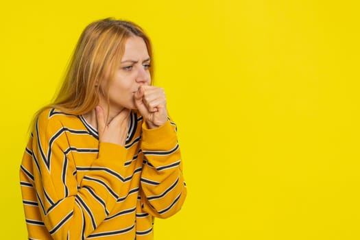 Unhealthy Caucasian blonde woman in blouse coughing with seasonal flu symptoms covering mouth with hand, feeling sick, allergy, fever or viral infection. Unwell ill girl isolated on yellow background