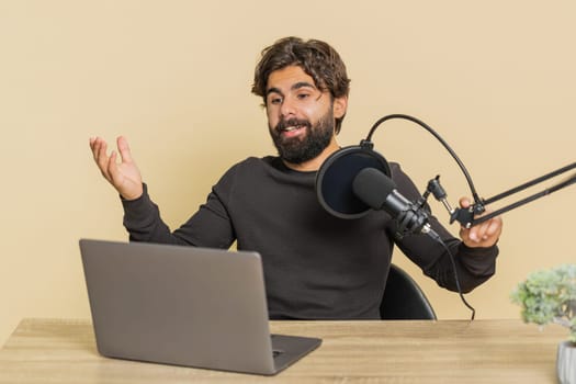 Happy blogger man recording podcast at table, giving workshop, webinar. Young Indian male speaking at microphone and laptop webcam, making video call. Handsome Hindu guy isolated on beige background.