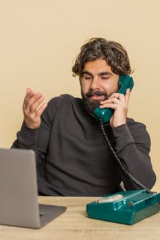 Happy bearded Hindu businessman sitting at table talking on wired telephone call conversation communication with client. Young freelancer Indian guy using laptop computer on beige studio background.