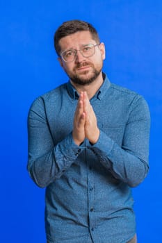 Please, God help. Caucasian middle-aged man praying, looking at camera and making wish, asking with hopeful imploring expression, begging apology, meditation. Adult guy isolated on blue background