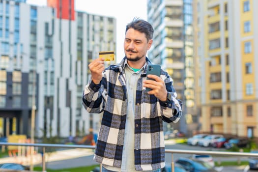 Caucasian mature man using credit card and smartphone while transferring money purchases online shopping order food delivery on internet outdoors. Guy standing against building on downtown city street