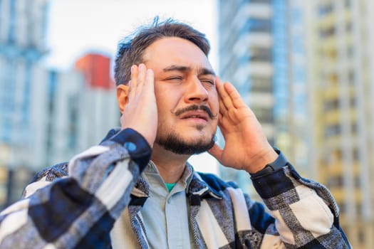 Sick mature Caucasian man feels headache on downtown city street. Middle-aged tourist guy massages her forehead and temples with his hands against building. Upset businessman suffering from migraine.