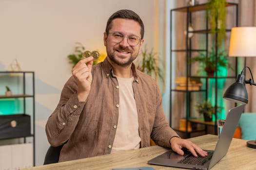 Young businessman freelancer stock trader earning holding showing bitcoins after online monitoring trading operations. Increasing wealth, financial prosperity. Guy with laptop at home office table.