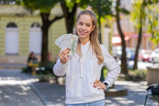 Portrait of successful rich Caucasian girl gambler gambling addiction business woman winner wins days holding fan of dollars, shows finances with thumbs up gesture receives loan salary on city street.