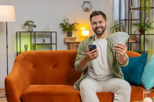Successful rich young Caucasian man holding and waving dollar bill money fan sitting on sofa couch at home. Happy adult guy winning online lottery game planning vacation calculating budget concept.