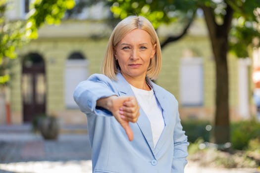 Sad upset disappointed Caucasian mature businesswoman female woman in formal suit showing thumb down sign gesture, rejection finger dislike negative reaction disagreement outdoors town city street.