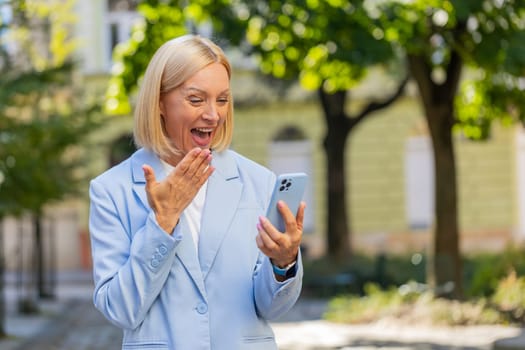 Happy shocked excited Caucasian mature businesswoman in formal suit mouth opened celebrate victory, achievement holding mobile phone good news win message rejoice business success outside city street.