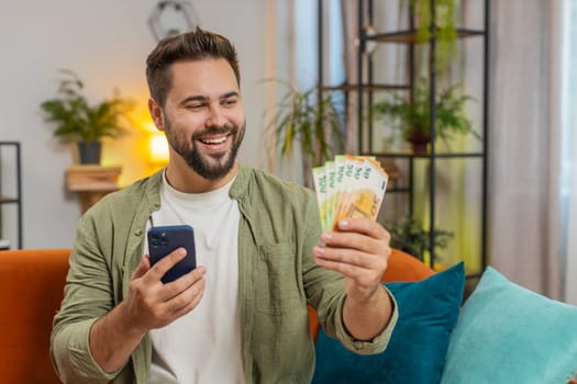 Successful rich young Caucasian man holding and waving euro bill money fan sitting on sofa couch at home. Happy adult guy winning online lottery game planning vacation calculating budget concept.