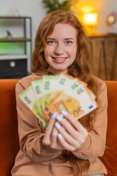 Successful rich young Caucasian woman holding and waving euro bill money fan sitting on sofa couch at home. Happy adult girl winning online lottery game planning vacation calculating budget concept.