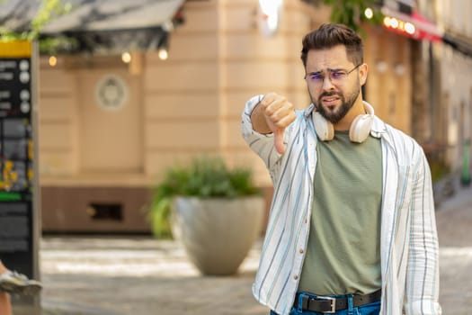 Dislike. Upset Caucasian young man showing thumbs down sign gesture, expressing discontent disapproval, dissatisfied bad work, mistake outdoors. Displeased guy tourist standing on urban city street.
