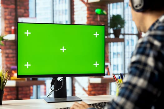 Worker listening music, analyzing data on green screen PC at desk, compiling analytical reports. Close up of employee wearing headphones using mockup computer, finishing project