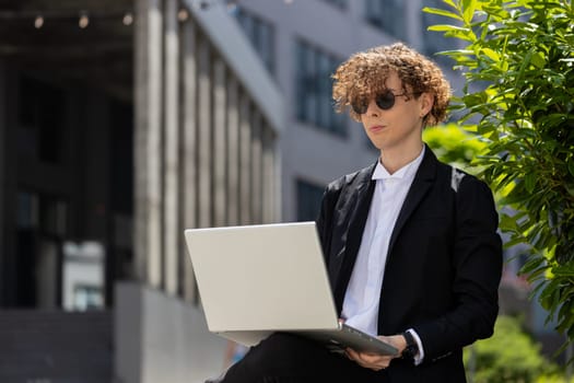 Caucasian young businessman investor employer entrepreneur sitting outside office building opening laptop and working on business project in city. Freelancer guy man in elegant suit and sunglasses.