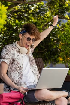 Handsome young man using laptop computer celebrating win good message news, lottery jackpot victory, giveaway online outdoors. Happy guy tourist with netbook sitting on bench in city. Town lifestyles.
