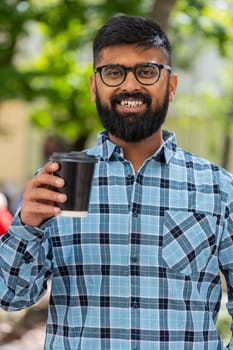 Happy Indian bearded man enjoying morning coffee hot drink and smiling outdoors. Relaxing, taking a break. Hispanic guy standing on urban city street drinking coffee to go. Town lifestyles. Vertical