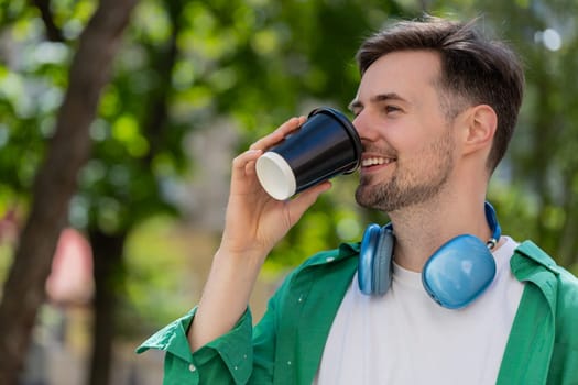 Happy Caucasian adult man enjoying morning coffee hot drink and smiling. Relaxing, taking a break. Young guy standing on urban city center street, drinking coffee to go. Town lifestyles outside.