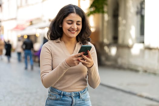 Indian young woman using smartphone typing text answering messages chatting online looking mobile screen social media app outdoors. Hispanic girl holding phone in urban city street. Town lifestyles
