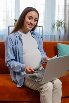 Happy pregnant woman counting money pressing keyboard keys on laptop smiling. Positive beautiful expectant lady sitting on sofa couch buying online at home indoors. Comfort and convenience. Vertical