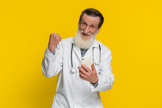 Happy senior doctor man use mobile smartphone typing browsing say Wow celebrating success victory winning lottery jackpot goal achievement play game, good positive news triumph on yellow background.