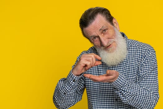 Need some more, please give me. Senior old man showing a little bit gesture with sceptic smile showing minimum sign measuring small size begging help. Elderly grandfather on yellow background indoors