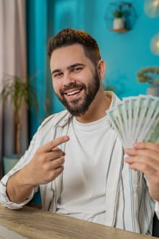 Young businessman guy holding fan of cash money dollar banknotes showing pointing on banknotes, celebrate, success career, online income, wealth at home office desk. Remote distant working. Vertical