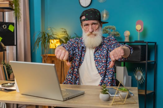 Dislike. Upset stylish senior man working on laptop computer at home office showing thumbs down sign gesture, expressing discontent, disapproval dissatisfied bad work. Displeased grandpa freelancer.