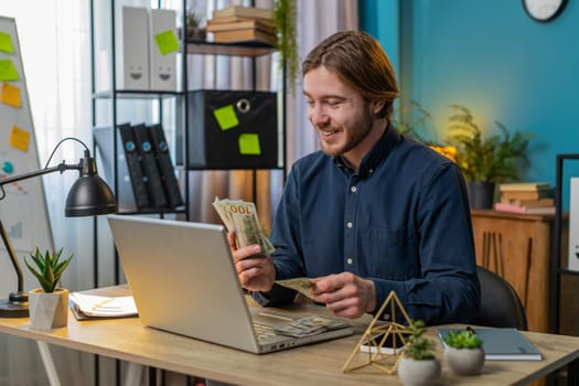 Excited businessman guy holding fan of cash money dollar banknotes showing thumbs up and pointing at laptop, celebrate, success career online income, wealth at home office desk. Remote distant working