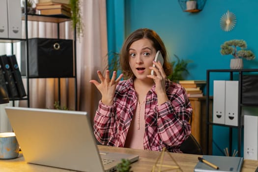Young businesswoman freelancer answering to colleague on mobile phone call, using smartphone and laptop. Conversation. Caucasian woman working online remote distance job, talking at home office desk.
