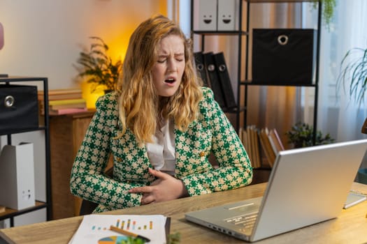 Disappointed Caucasian freelancer feeling overeating during work process on laptop in home office. Upset businesswoman freelancer suffering from painful stomach ache, period cramps, menstrual pain.