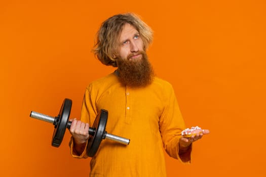 Caucasian man holding dumbbell training exercising looking at sweet donut cake with desire choosing between fitness sport diet vitamin nutrition or junk food. Redhead guy isolated on orange background