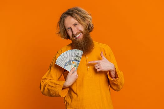 Rich pleased happy Caucasian man waving money dollar cash banknotes bills like a fan, success business career, lottery winner, big income wealth. Young adult guy isolated on orange background indoors