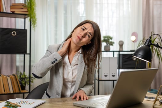 Tired Caucasian young businesswoman suffers from spine neck muscle pain at home office workplace working on laptop. Overworking. Freelancer girl in suit attempts to relieve pain by massaging at table.