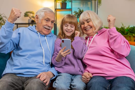 Surprise Caucasian grandfather, grandmother and granddaughter using smartphone while celebrating success at home. Happy girl with grandparents amazed by online game win sitting on couch in living room