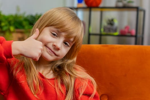 Like. Happy young Caucasian child girl looking approvingly at camera showing thumbs up like sign positive something good, positive feedback. Blonde teenager kid at home in living room sits on sofa