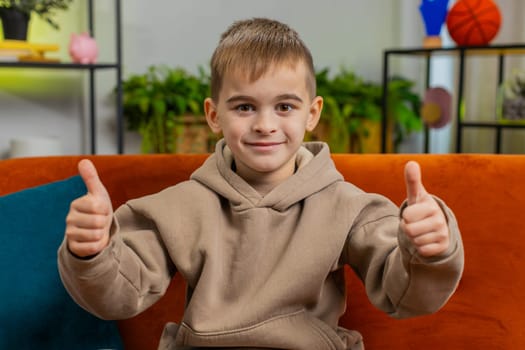 Like. Happy young Caucasian child boy looking approvingly at camera showing thumbs up like sign positive something good, positive feedback. Male little teenager kid at home in living room sits on sofa
