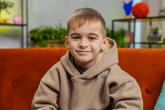 Close-up portrait of happy smiling cheerful Caucasian preteen school boy 8 years old. Young lovely blonde little child kid looking at camera at home play living room apartment sitting on orange sofa