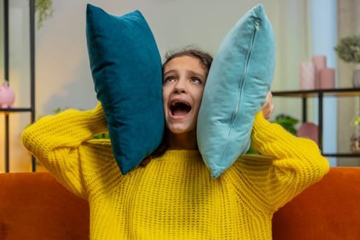 Repair work at neighbors. Irritated child girl kid shouting cover ears with pillows annoyed by noisy loud music suffers headache wishes silence. Thin walls at home flat without sound insulation