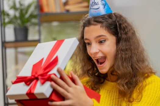 Happy 15 years old child girl wears festive birthday cap hat hold gift box with ribbon congratulating. Teenager female kid celebrating party event opening delivery greeting present at home room