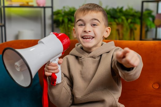 Preteen child boy talking with megaphone proclaiming news, loudly announcing advertisement warning using loudspeaker to shout speech. Male kid at home room apartment on sofa, pointing finger to camera