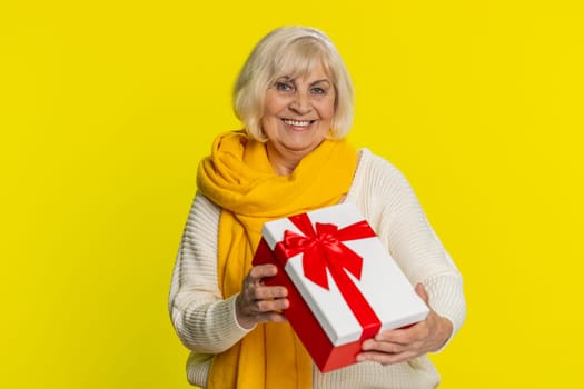 Positive smiling senior woman presenting birthday gift box stretches out hands, offer wrapped present career bonus, celebrating party, promotion discount sale. Elderly grandmother on yellow background
