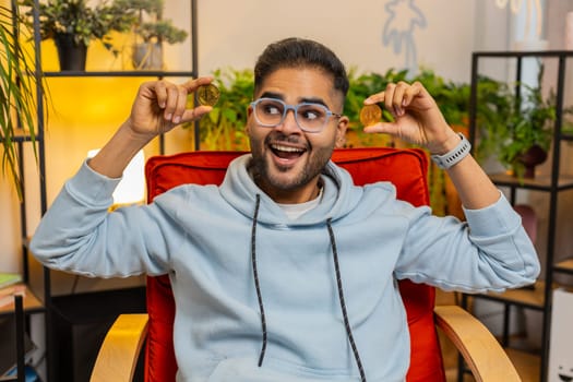 Indian man freelancer holding two gold coins, successful developer programmer. Arab guy stock trader earning bitcoins after online monitoring trading operations. Increasing wealth financial prosperity