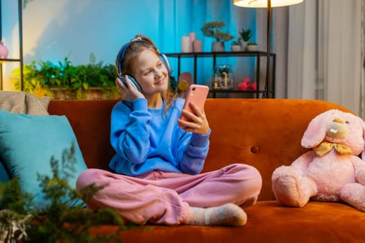Happy overjoyed teenager girl kid in wireless headphones relaxing sits on couch at evening home room choosing listening favorite energetic disco dancing music. Children weekend leisure activities