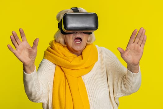 Excited happy senior woman using headset helmet app to play simulation game. Watching virtual reality 3D 360 video. Old grandmother in VR goggles isolated on yellow wall background. Future technology